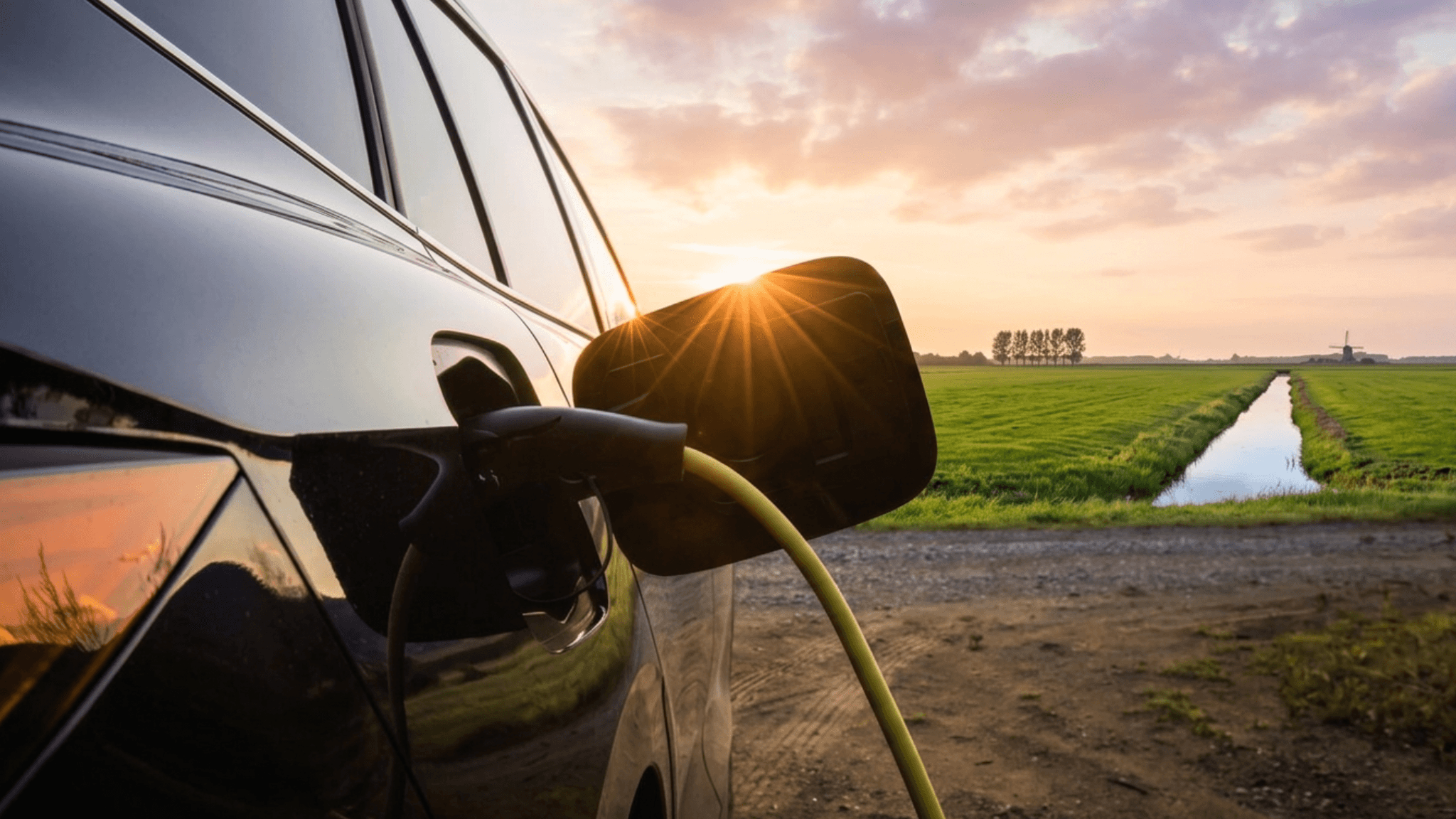 Elektrische auto staat aangesloten op een laadpunt langs een landelijke weg, met weilanden en een watergang op de achtergrond.