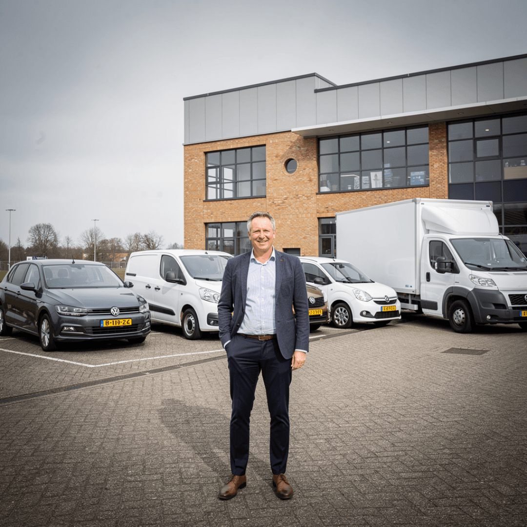 Persoon staat op een parkeerterrein voor een modern bedrijfspand, omringd door personenauto's en bedrijfswagens