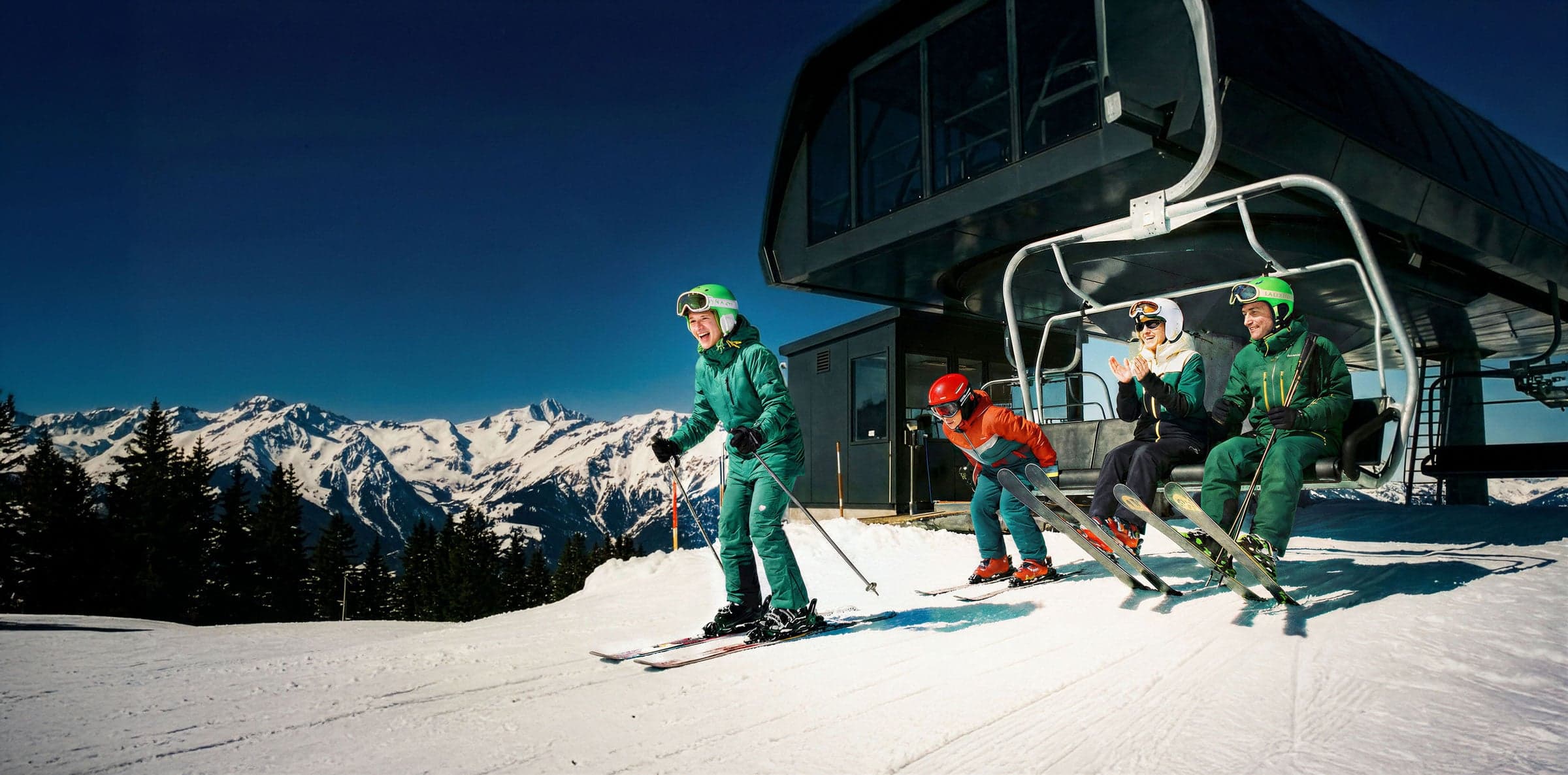 Meerdere personen stappen uit een skilift bovenaan een skipiste, met besneeuwde bergtoppen op de achtergrond
